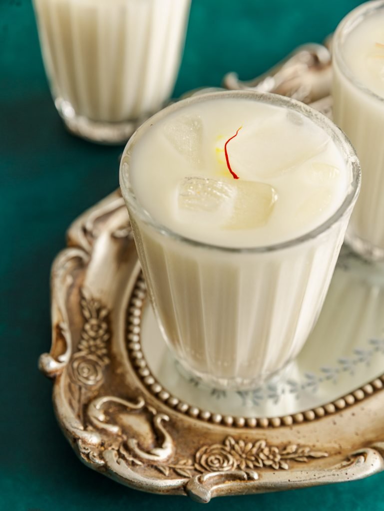 Mexikanische Safran-Kardamom-Horchata in einem Glas mit Eiswürfeln, dekoriert mit Zimtstange und goldenen Safranfäden – cremiger, erfrischender Reisdrink.